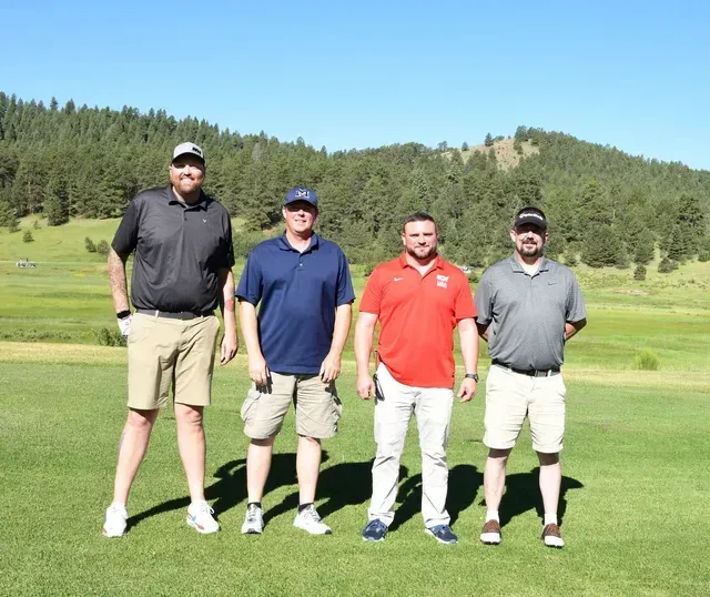 Four men standing on a golf course, posing for a photo. Sunny day. Mountains in the background.