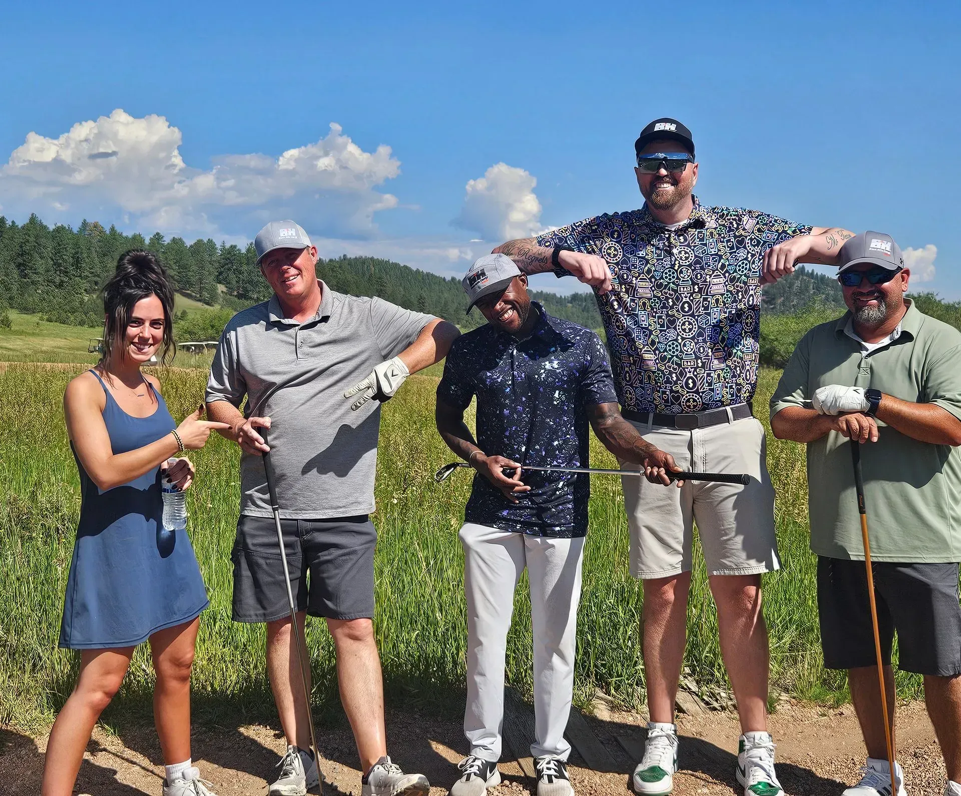 Five people pose with golf clubs outdoors: woman in blue dress, men in golf attire with arms outstretched.