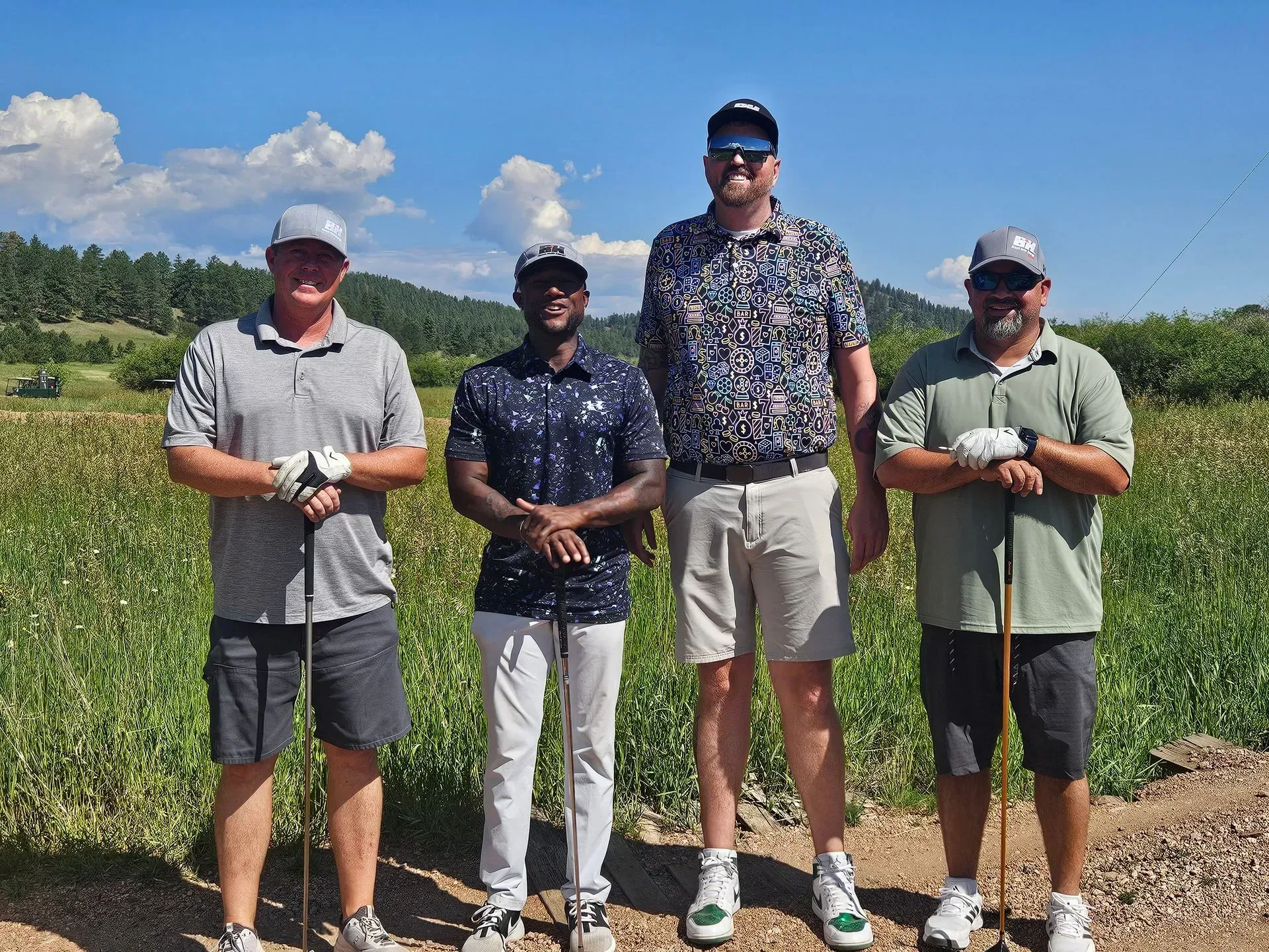 Four men in golf attire, standing on a dirt path in a grassy field, smiling, mountains visible in background.