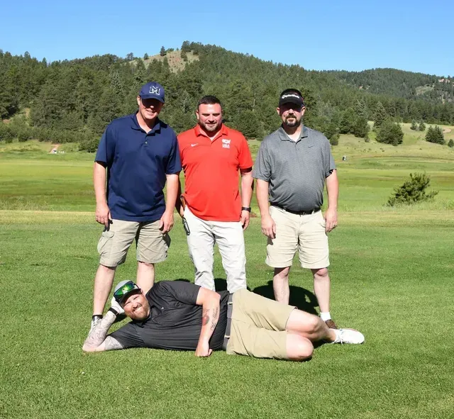 Four men on a golf course. Three standing, one lying on the ground. Green grass, blue sky, and a mountain backdrop.