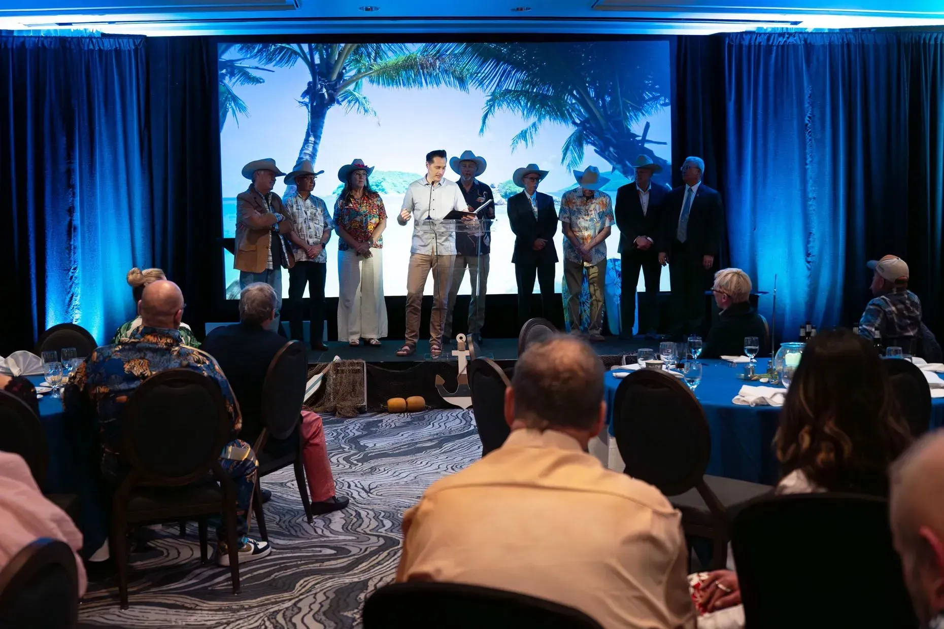 People on stage with a presenter at an event with a beach scene backdrop; attendees seated at tables.