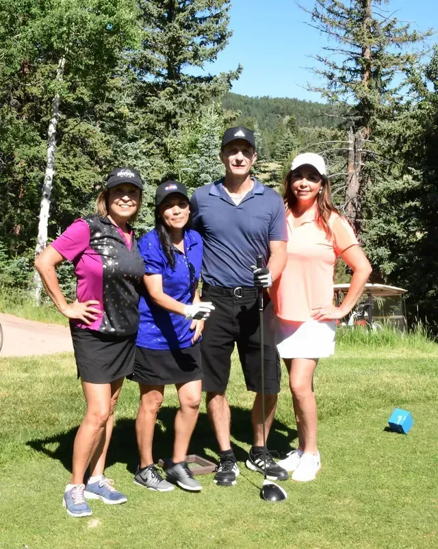 Four people pose on a golf course. Two women in skorts, two men in golf attire. Green grass, trees, sunny day.