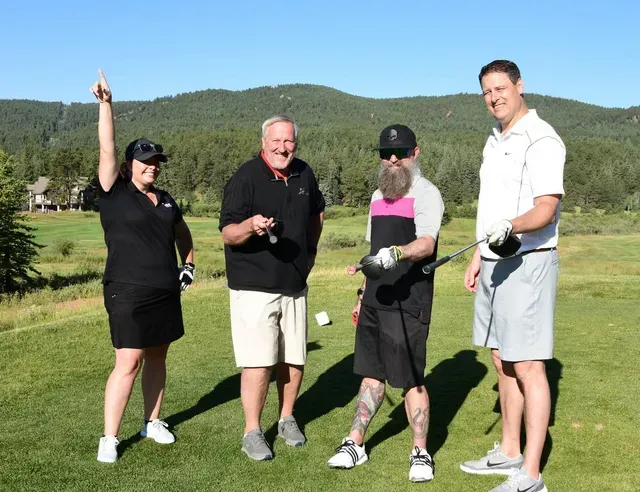 Four people on a golf course, one woman with arm raised, others holding clubs. Mountains in background.