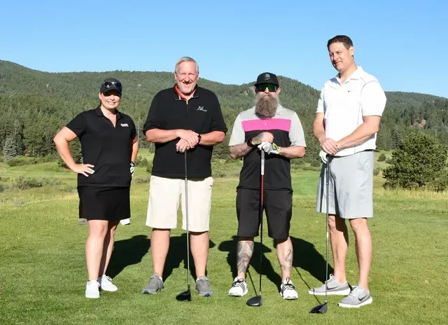 Four people on a golf course, smiling, holding clubs. Green grass, mountains in the background.