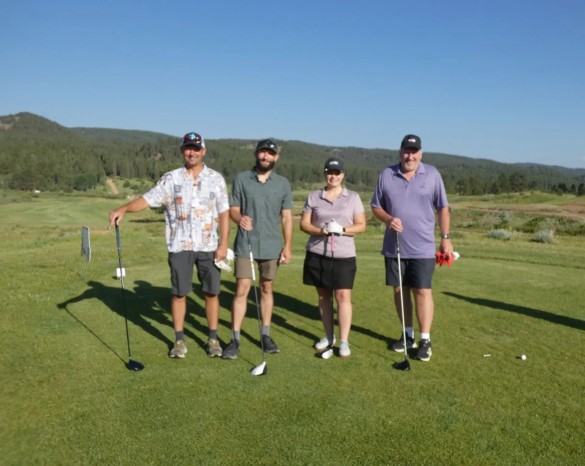 Four golfers pose on a green, sunny day. They stand holding clubs, smiling, with mountains in the background.