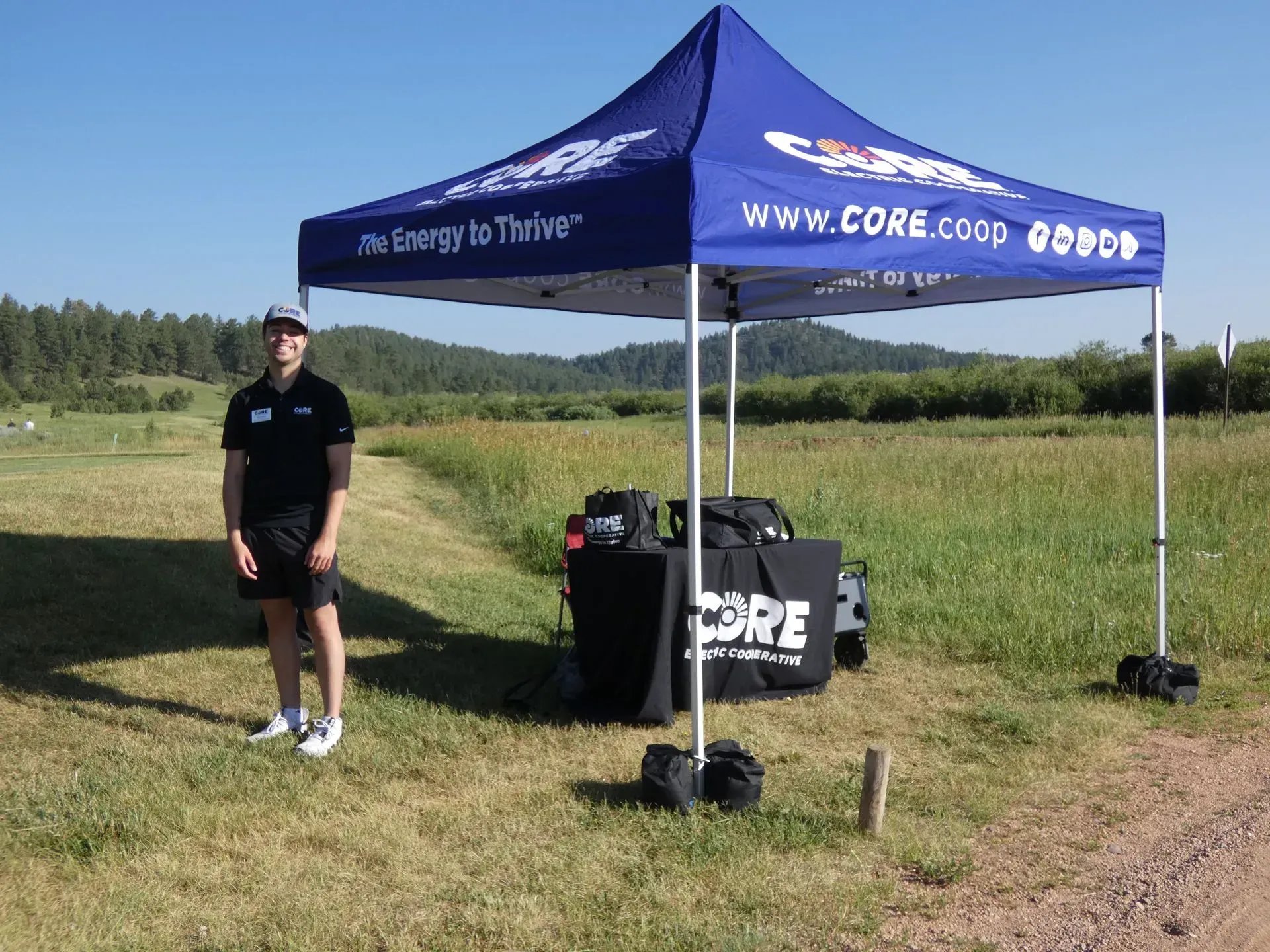 Man stands by CORE booth in field, wearing black shirt and shorts. Blue canopy, sunny day.