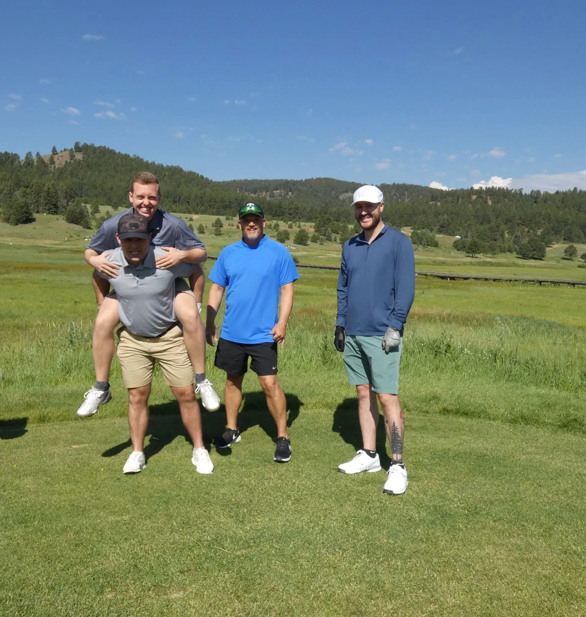Four men on a golf course. One gives another a piggyback ride. Two others stand nearby. Blue sky and green landscape.