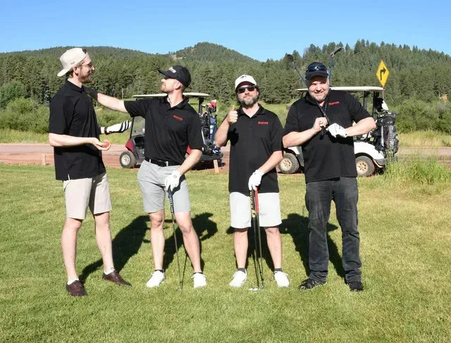 Four men pose on a golf course. They wear black shirts, some with hats, holding clubs, with golf carts in the background.