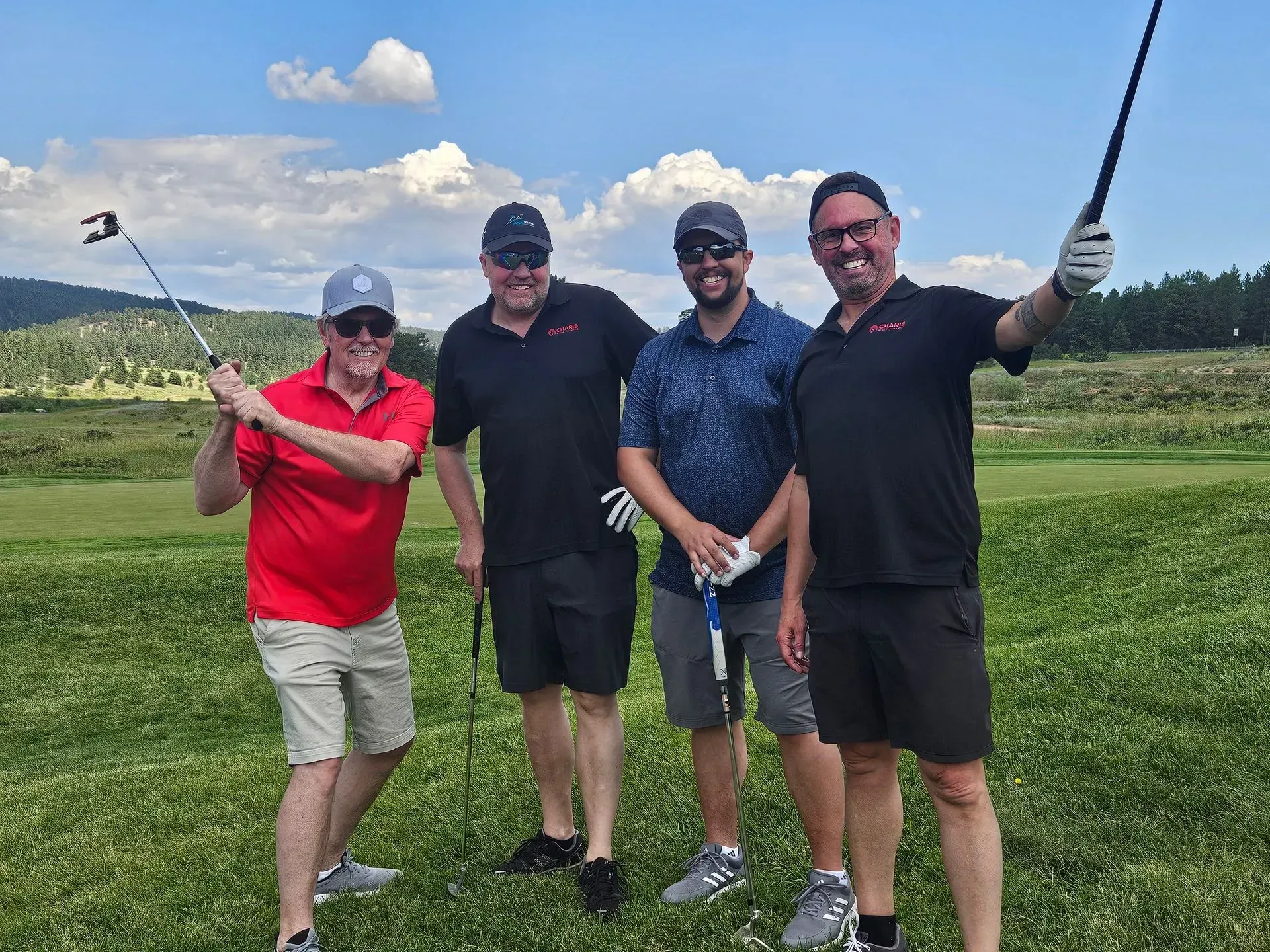 Four men on a golf course, one swinging a club. Smiling in the sun with green grass and blue sky.