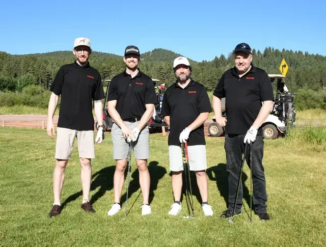 Four men in black shirts and golf attire pose on a grassy course with golf carts in the background.