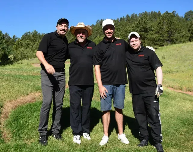 Four men pose on a golf course, smiling. They wear black shirts, hats, and golf attire.