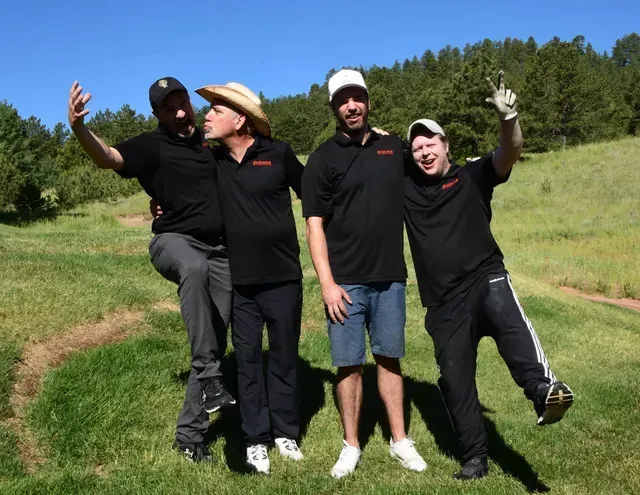 Four men on a golf course posing for photo: one with arm up, another making a face, two others smiling.