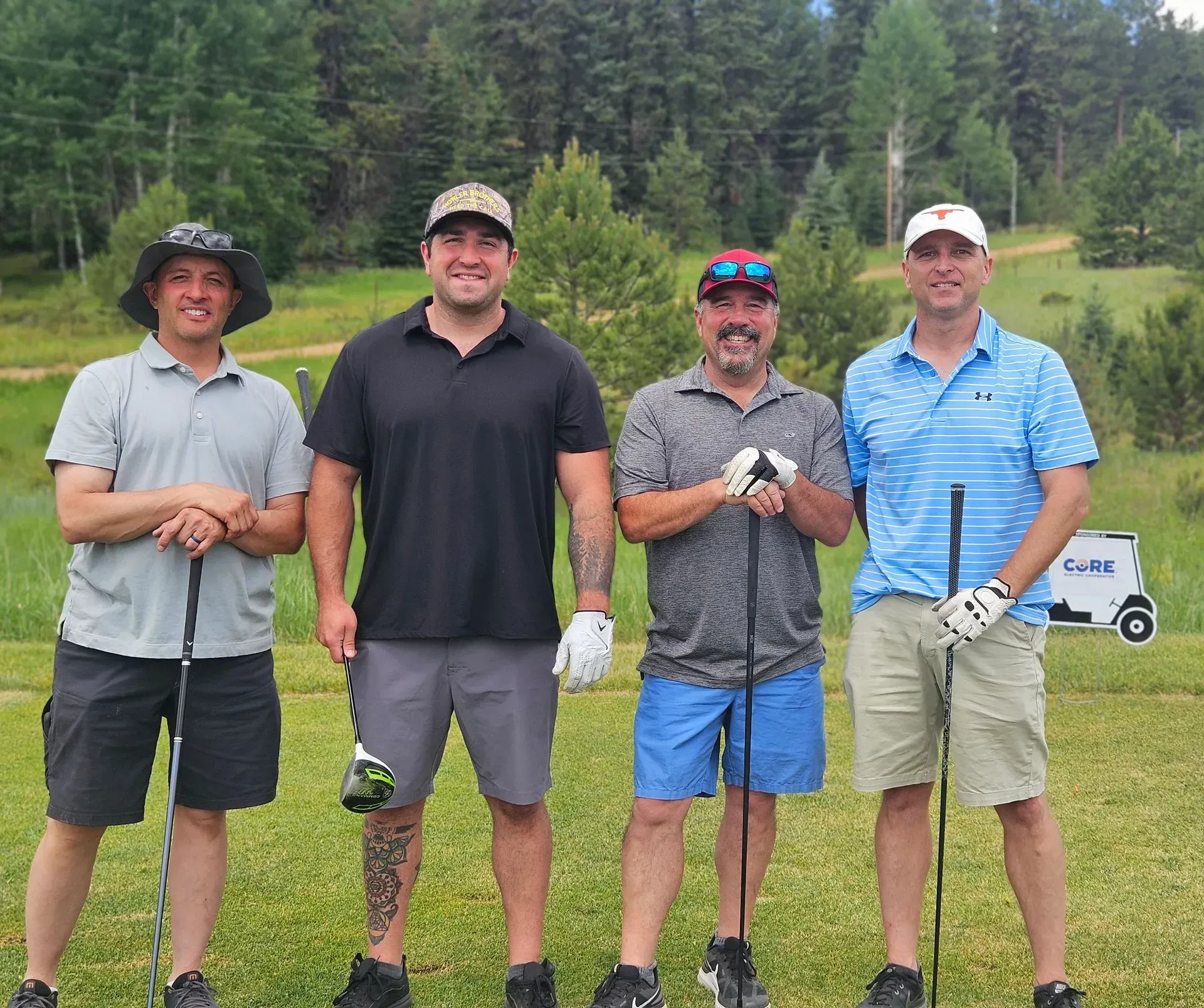 Four men smiling, posing with golf clubs on a green course, with trees in the background.