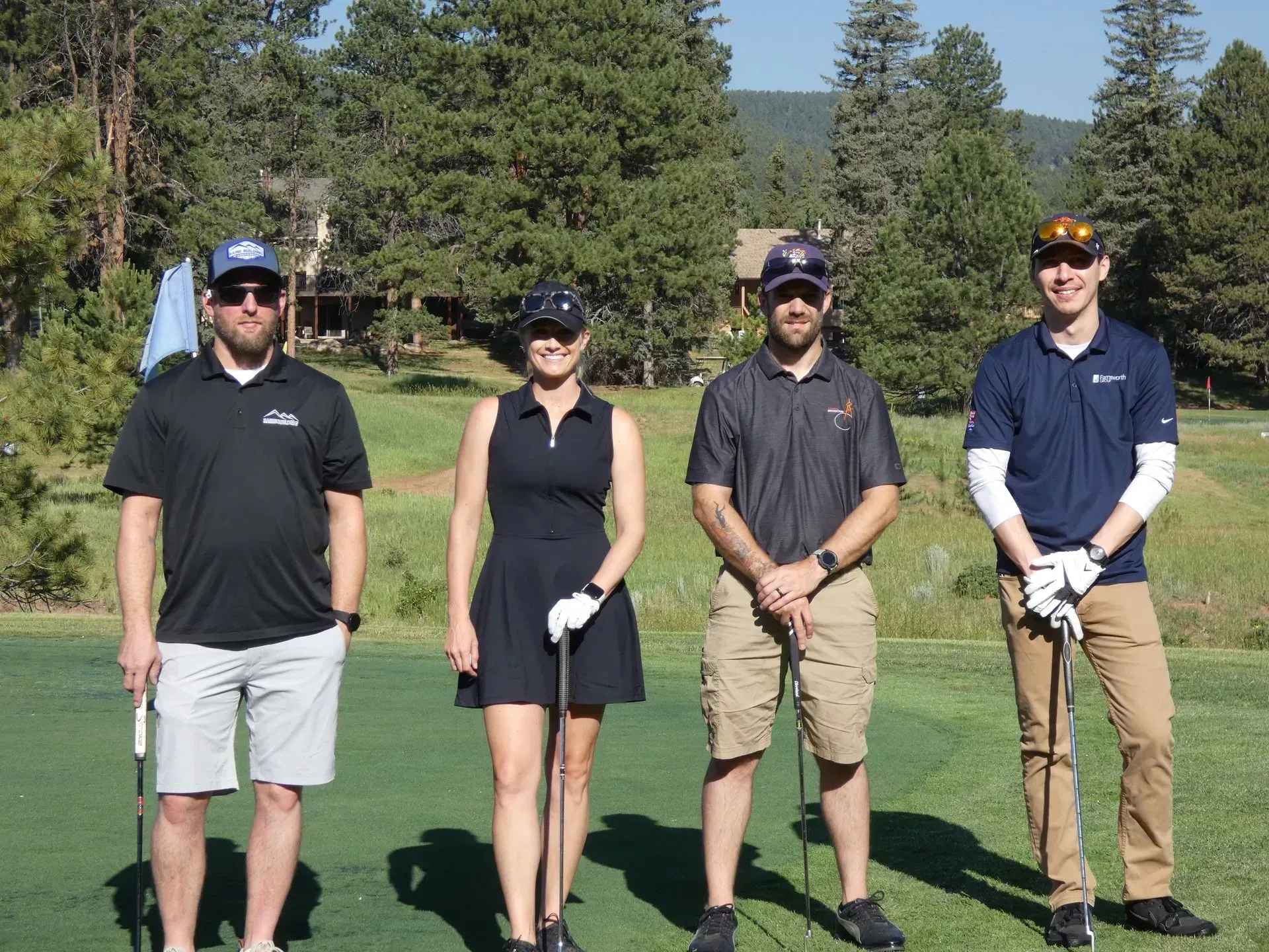 Four people stand on a golf course, holding clubs. One woman in a dark dress, three men in casual attire. Green grass, trees, sunny.