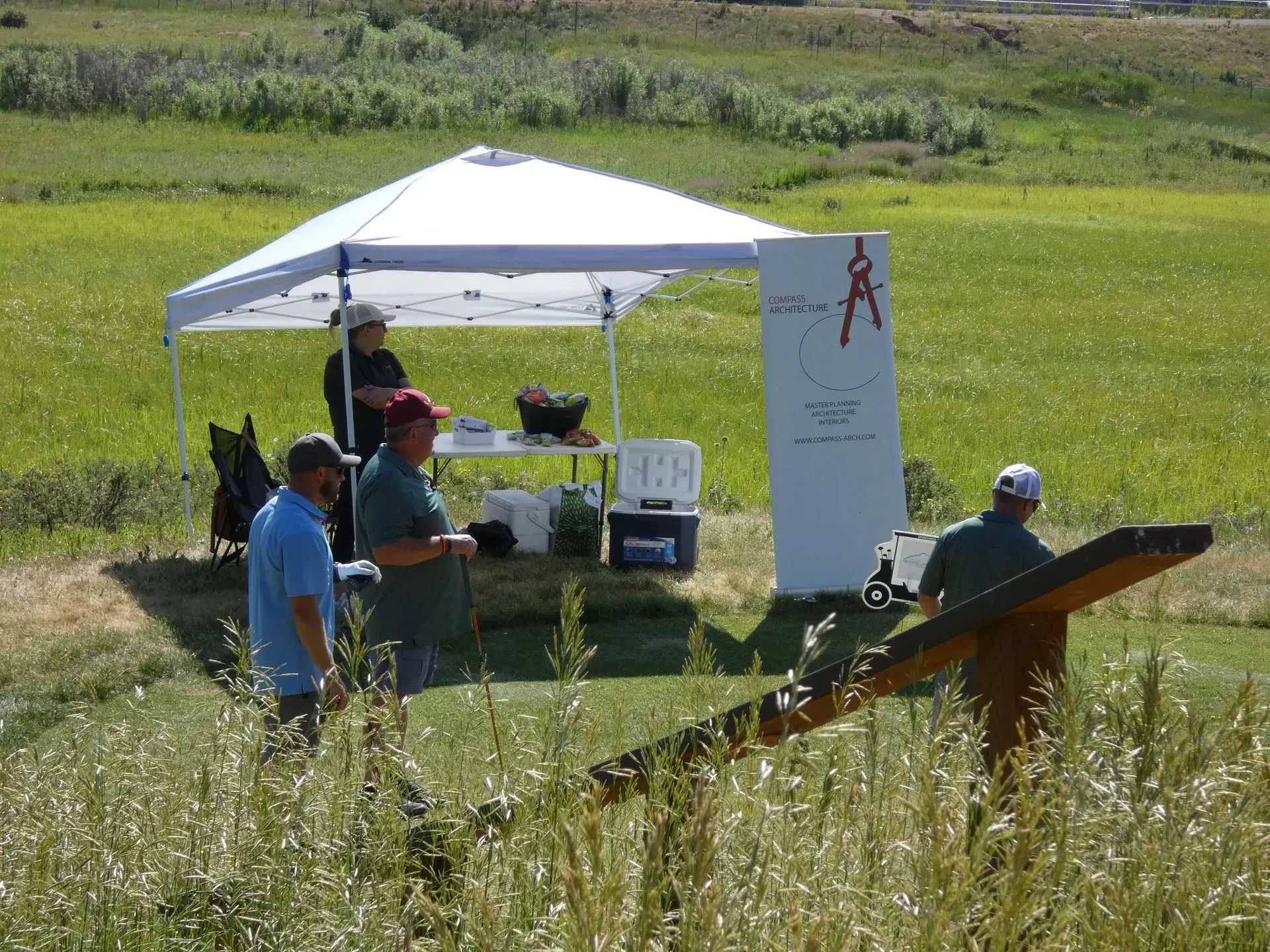 Golfers at a refreshment stand on a sunny day. White tent with logo, cooler, and people. Green field and sky.