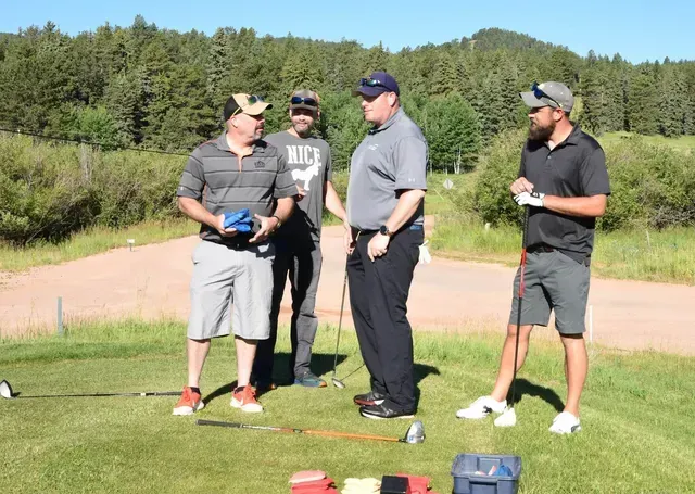 Four men on a golf course, talking. One holds a blue item, all wearing casual attire in sunny weather.