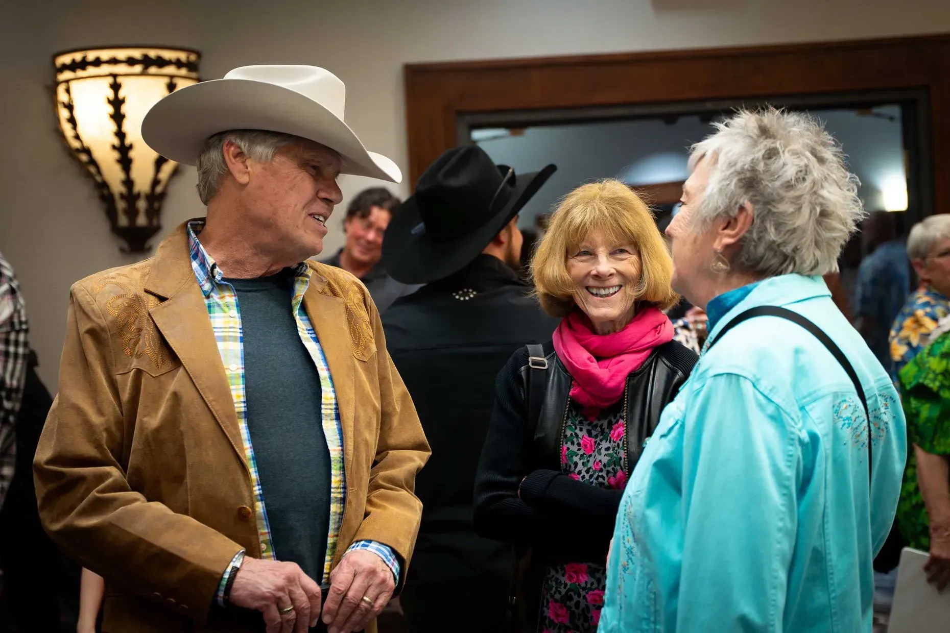 Group of people socializing indoors; man in cowboy hat and jacket, woman in pink scarf smiling.