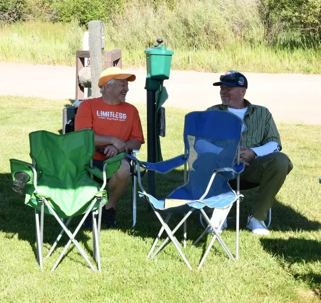 Two men sit in folding chairs outside, talking. One wears orange shirt, other wears hat and patterned shirt.