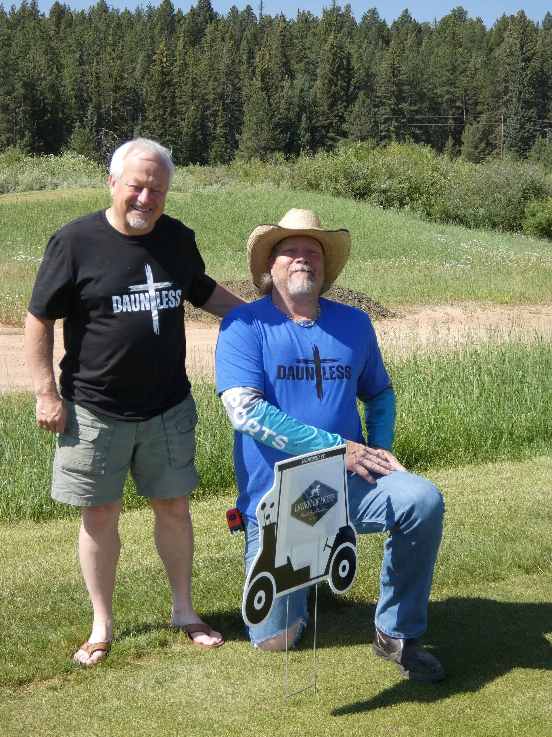 Two men outdoors; one kneels beside a sign, both wearing