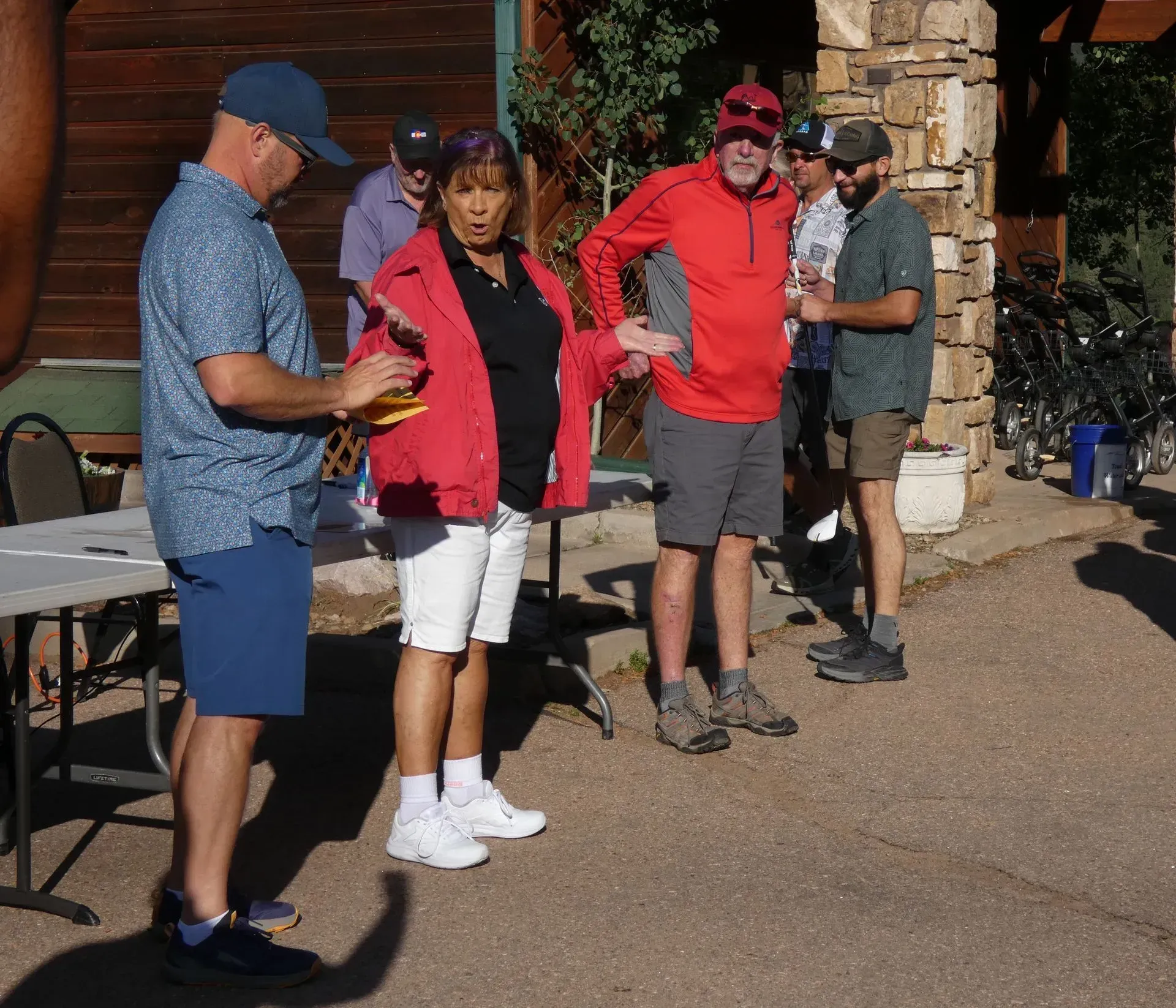 People outdoors near a building, some wearing hats and shorts. A woman in a red jacket, looks surprised.
