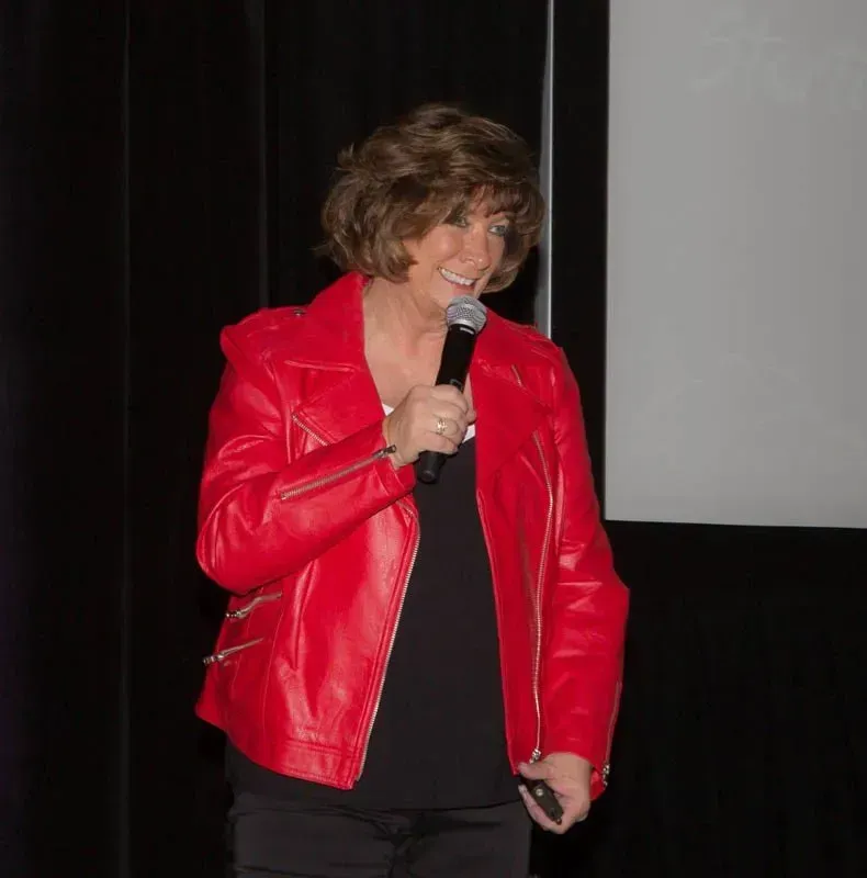 Woman in red leather jacket speaking into a microphone, standing in front of a screen.