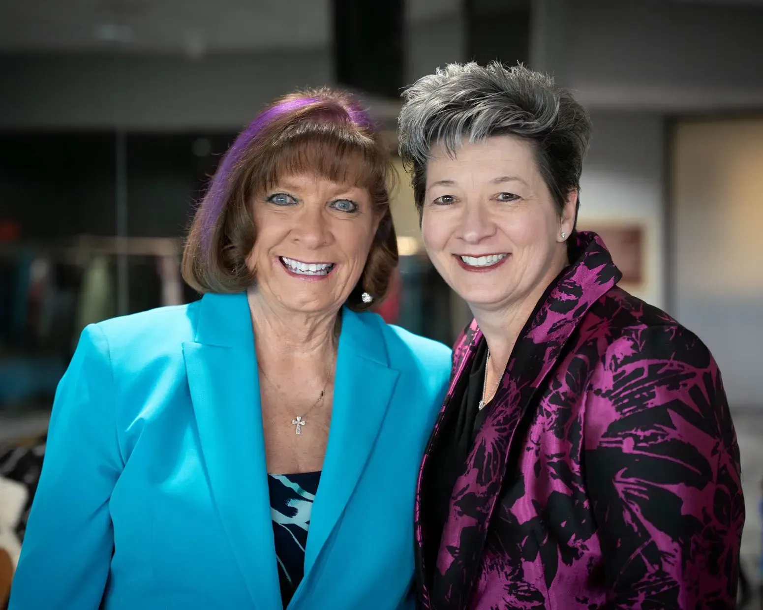 Two women smiling, one in blue blazer, one in magenta jacket, indoors.