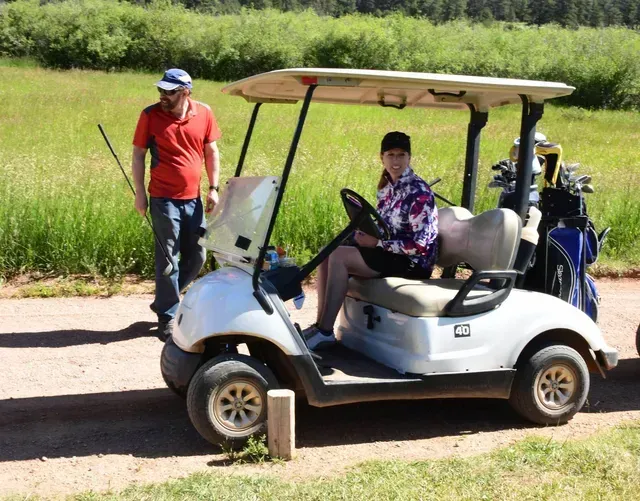 Woman driving a white golf cart, man standing by, on a golf course. Sunny day.