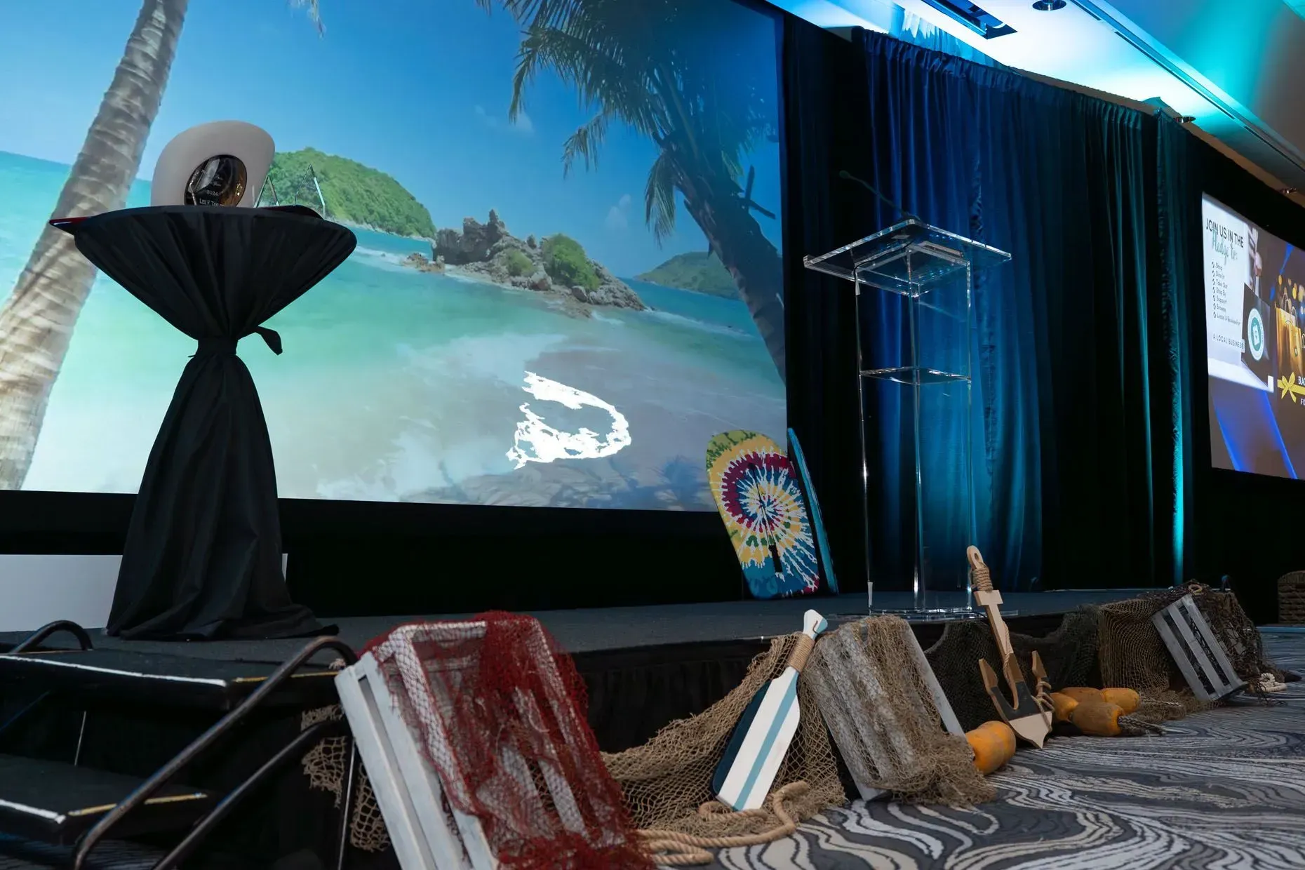 Stage set with a tropical beach backdrop, decorations include surfboards, netting, and a hat on a table.
