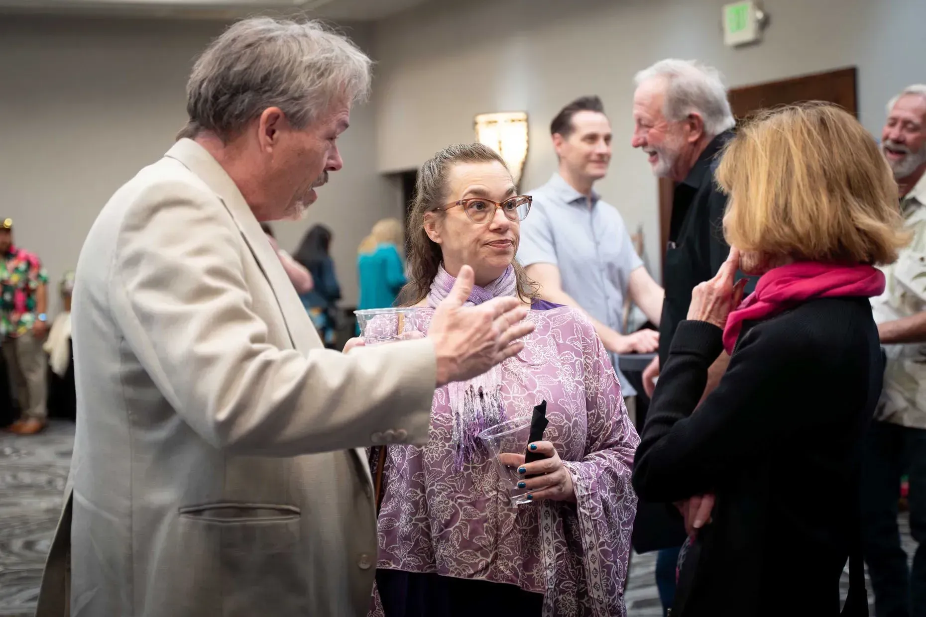 Group of people in conversation in a room; man gesturing, woman with glasses, and woman in a pink scarf.
