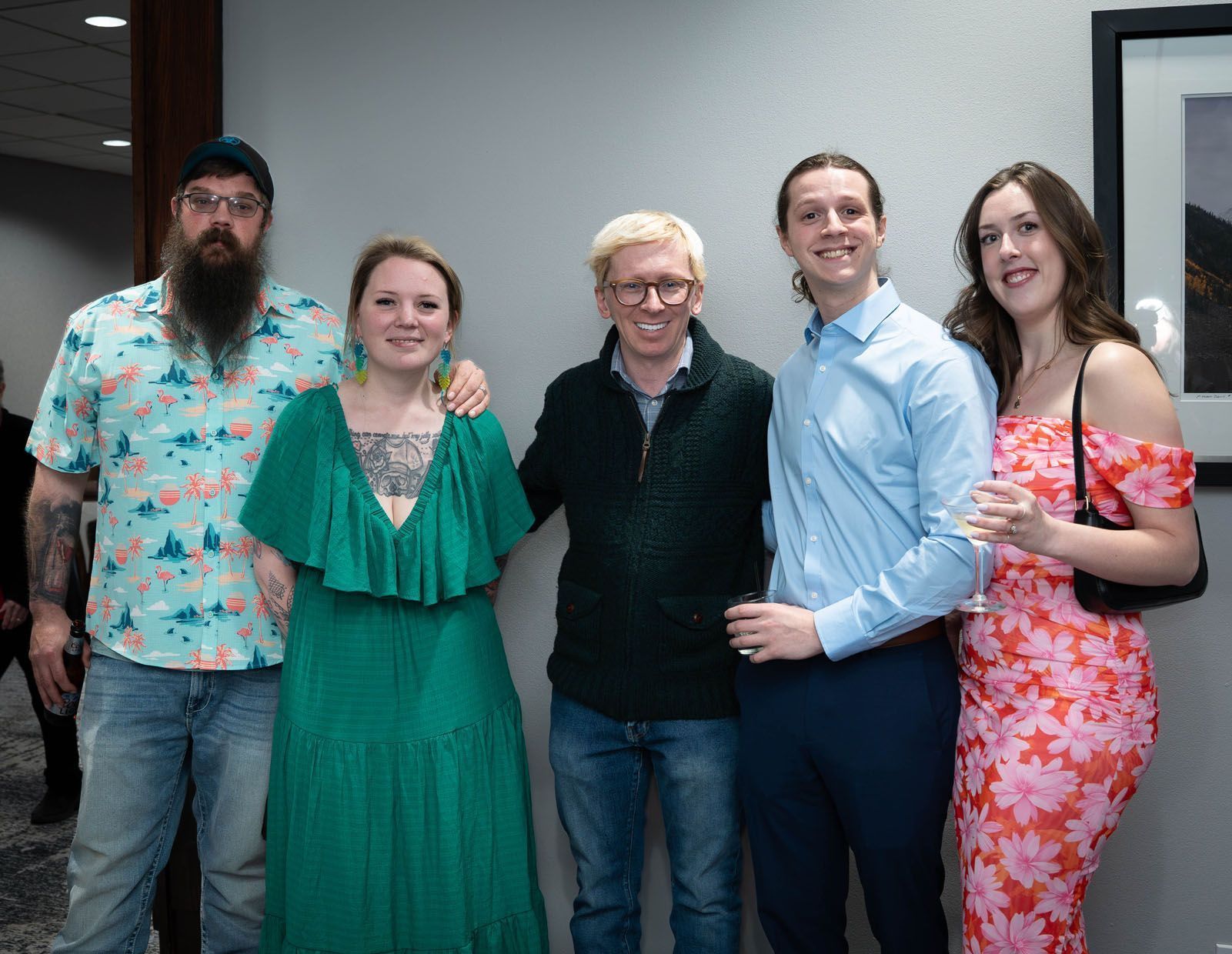 Five people smiling, posed in front of a wall. A man with a beard and floral shirt, a woman in green dress, another man in glasses and a green sweater, a man in a blue shirt, and a woman in a floral dress.