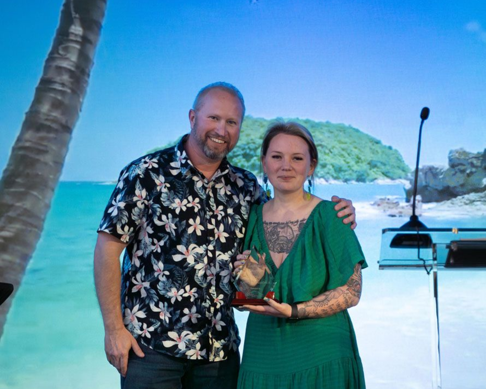Woman accepting an award from a man on stage. She wears a floral dress, he wears a blazer and jeans. Blue backdrop.