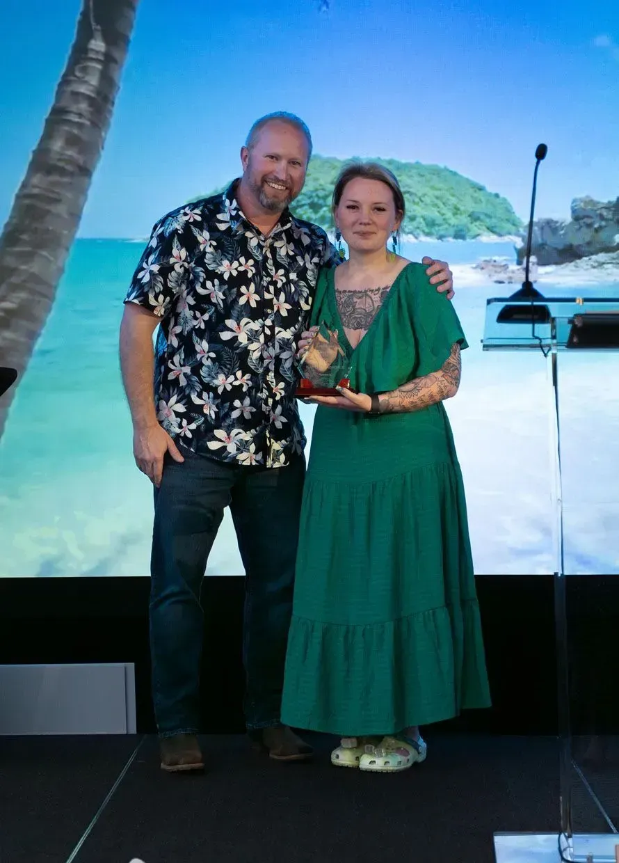 Man in Hawaiian shirt and woman in green dress smiling, holding trophy. Stage backdrop with tropical scene.