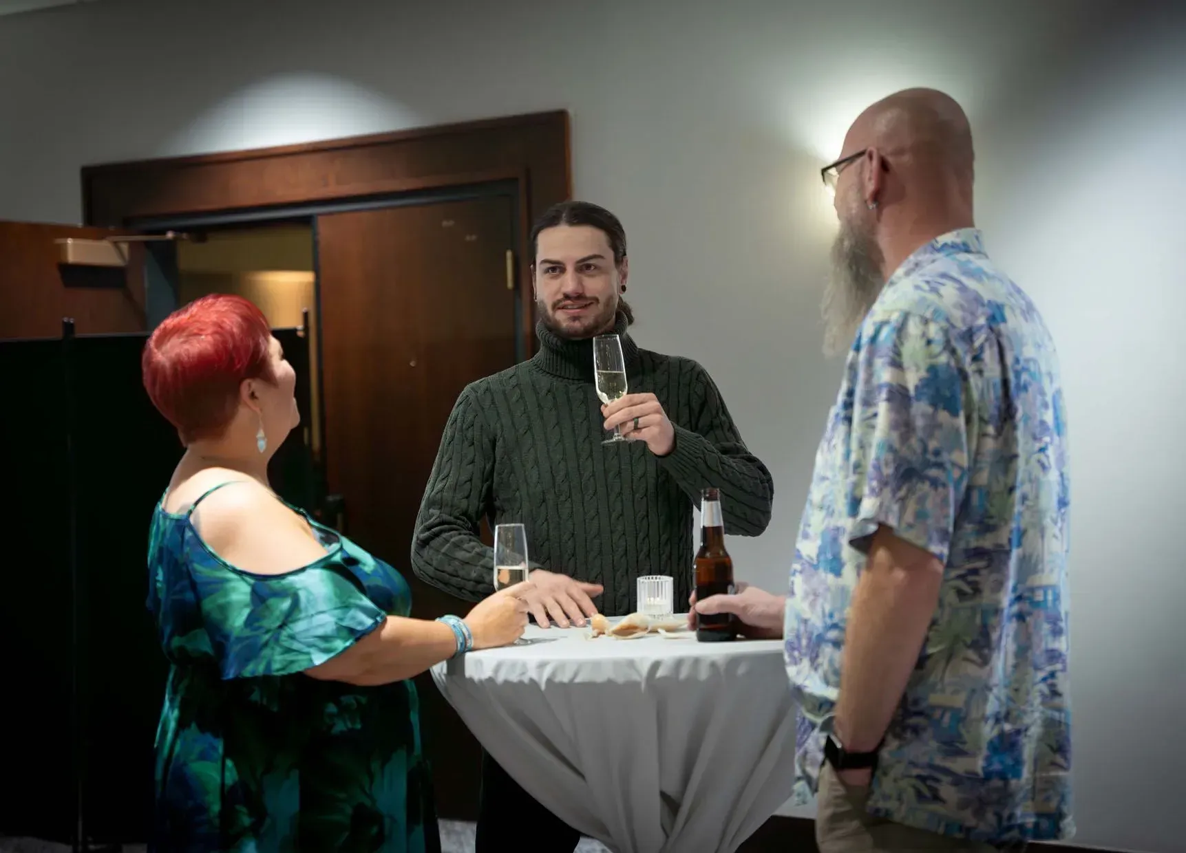Three people at a small table in a room, two holding drinks, all smiling and talking.
