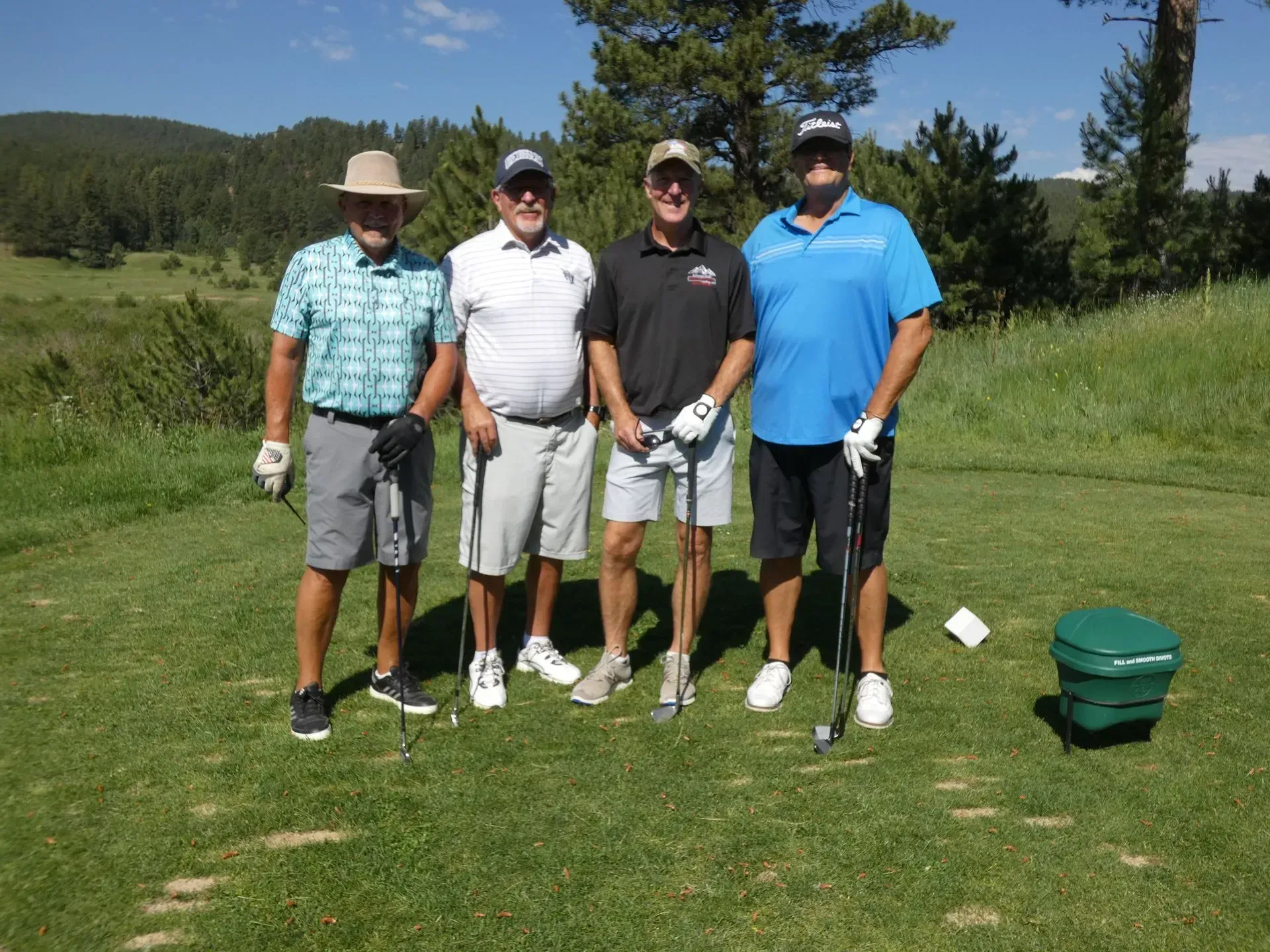 Four men on a golf course. They are standing on the tee, holding golf clubs, and smiling. Green grass and trees are in the background.