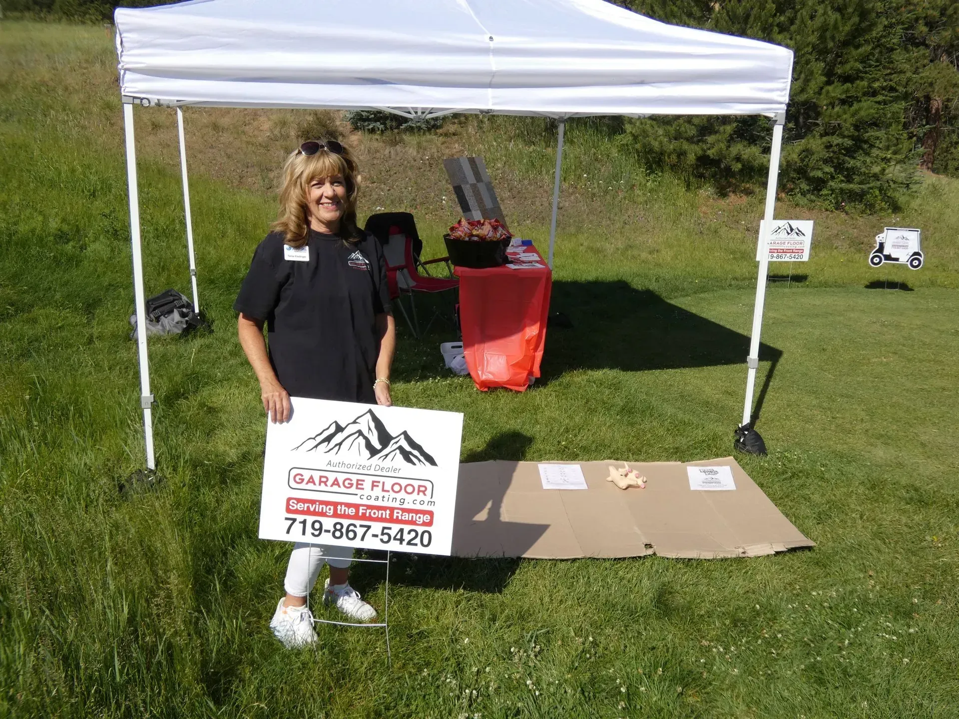 Woman at a booth in a grassy field. She holds a sign for
