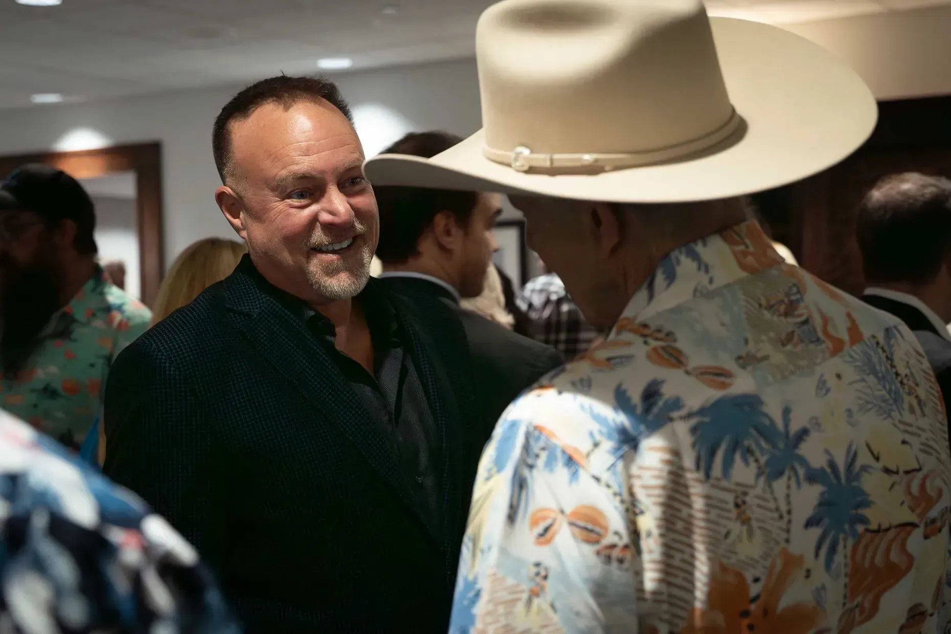 Man in black smiling at man wearing a cowboy hat and Hawaiian shirt, in a gathering.