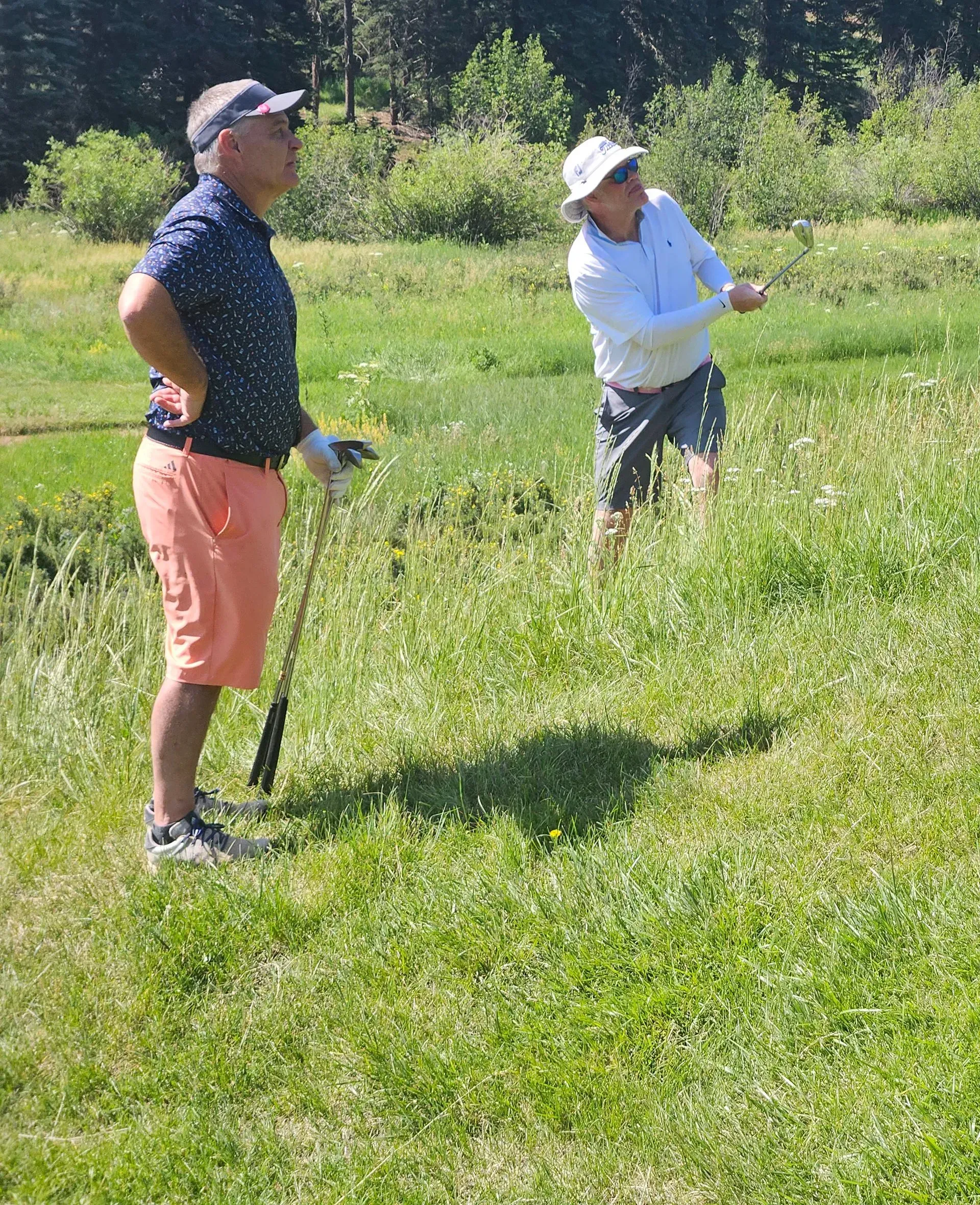 Two men golfing on a grassy course; one swings a club, the other watches.