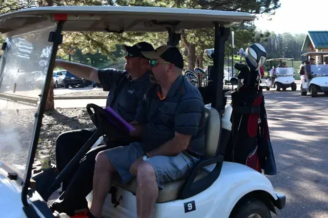 Two men in a golf cart, one pointing, another looking. Golf course in background.