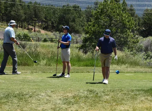 Three men golfing on a green, sunny day. One swings a club; others watch. Mountains in the background.