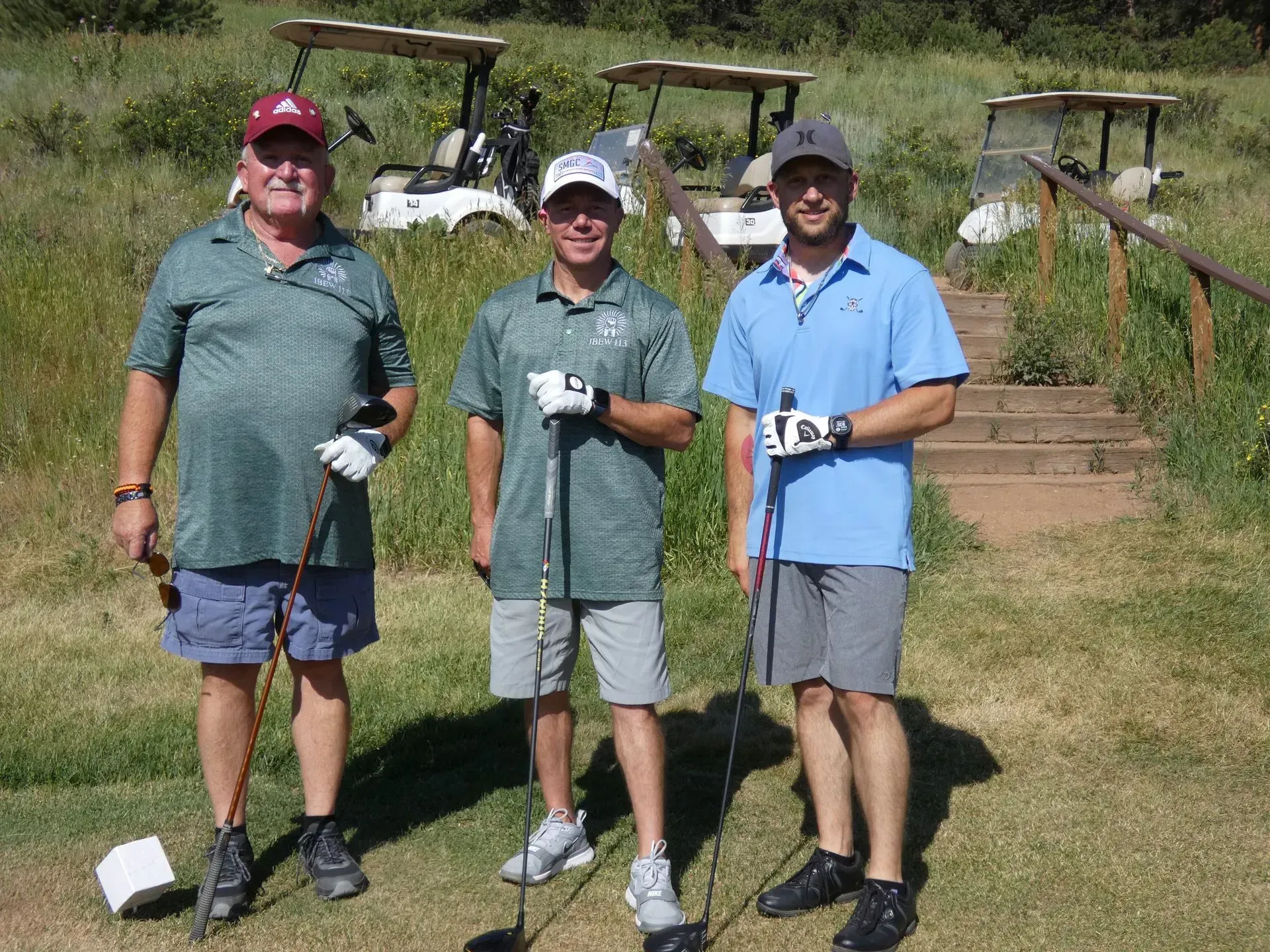 Three men on a golf course; two in golf attire, one in shorts; golf carts in the background, green grass and sunny day.