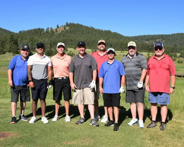 Nine men standing on a golf course, smiling, some wearing hats. Green grass, blue sky, trees in the background.