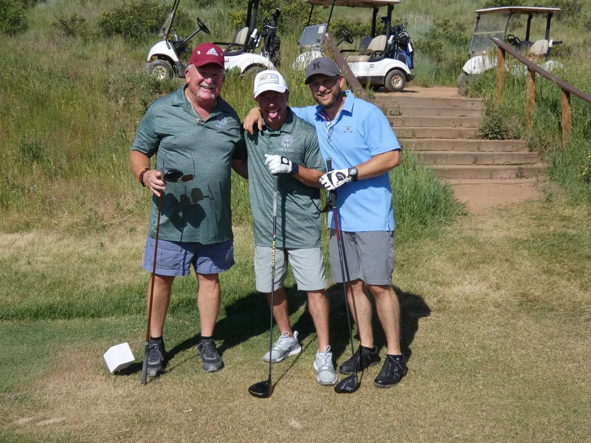 Three men smiling on a golf course, posing with clubs, golf carts, and steps in the background.