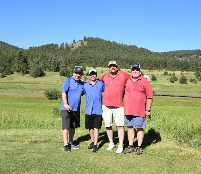 Four people stand on a grassy field with a mountainous backdrop; all are smiling, wearing caps and golf attire.