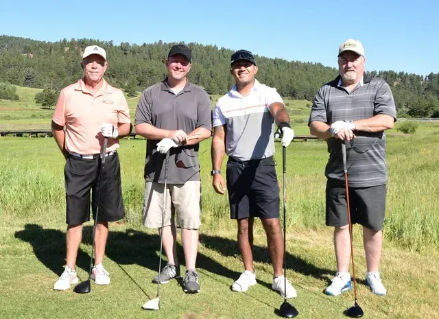 Four men pose on a golf course, holding clubs. They wear casual clothing; a green field and trees are in the background.