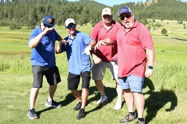 Four men on a golf course pose with silly faces; one covers his eye, another sticks out his tongue.