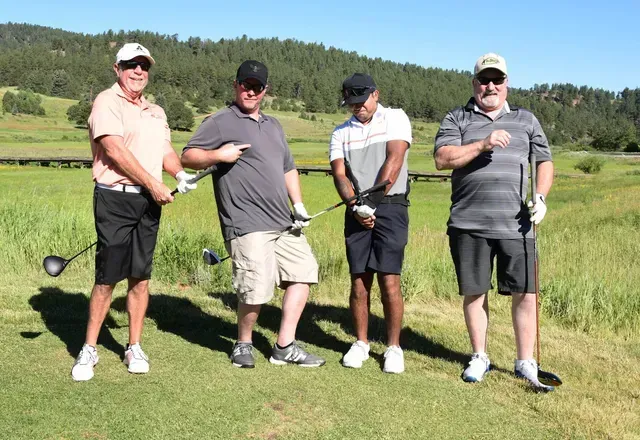 Four men on a golf course, posing with clubs. They wear casual clothes and hats, in front of a green landscape.