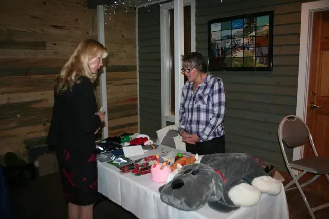 Two women at a table with gifts for sale; one is looking at the items, the other is standing.