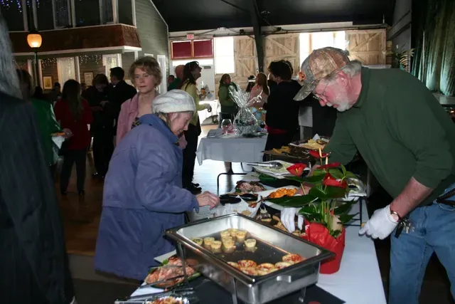 People at a buffet, one serving food wearing gloves, others in line at a brightly lit indoor event.