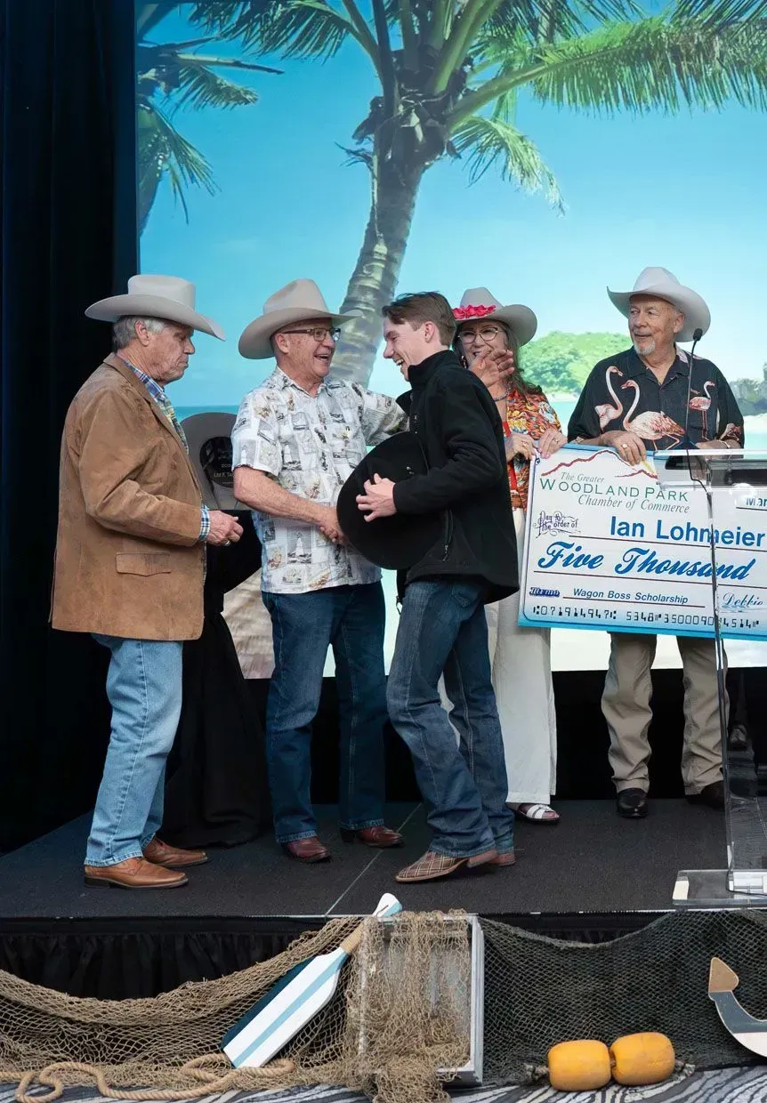Men in cowboy hats present a large check to a young man on a stage with a tropical backdrop.