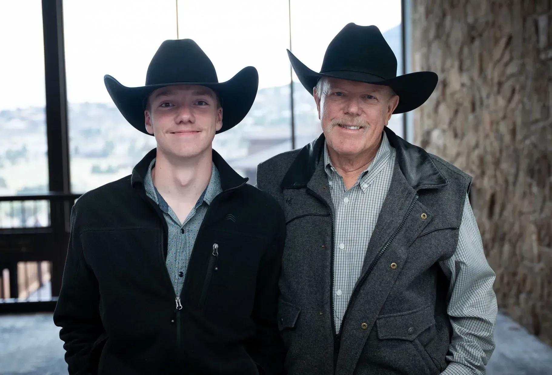 Two men, one younger, one older, wearing cowboy hats and jackets indoors.